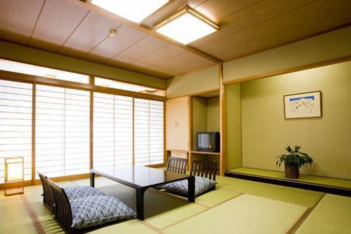 A tatami mat room with a low table and cushions, a television on a shelf, and a plant on the wall.