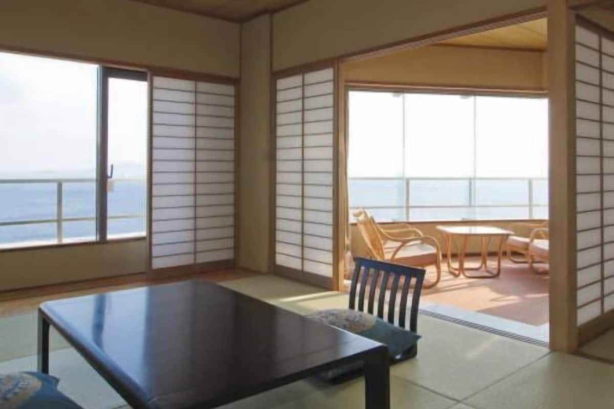 A room with tatami flooring, a low table, and sliding doors leading to a balcony with a view of the sea.