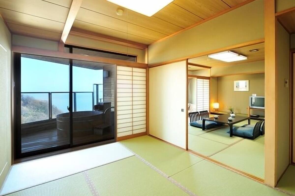 A traditional Japanese room with tatami flooring, a sliding door, a balcony with a view, and a small seating area with a table and chairs.