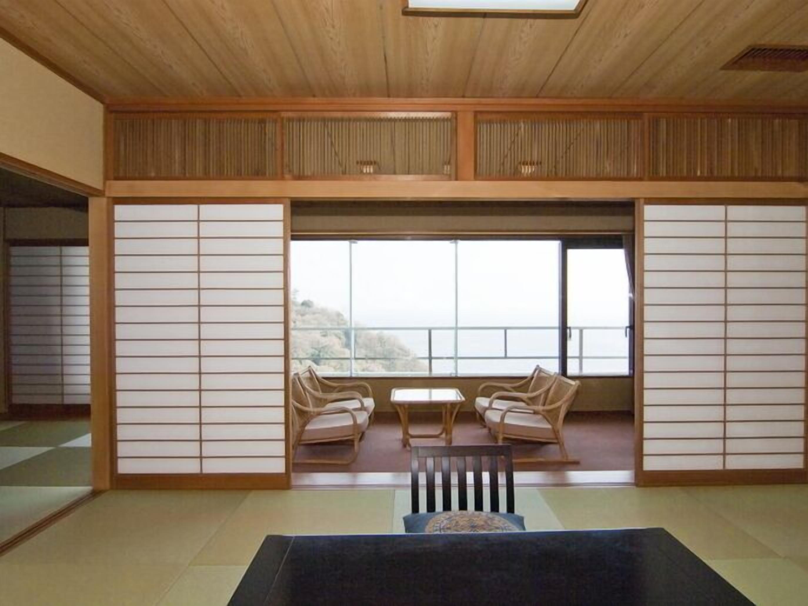 Una habitación japonesa tradicional con suelo de tatami, una mesa baja, dos sillas y vistas amplias a través de ventanas grandes.