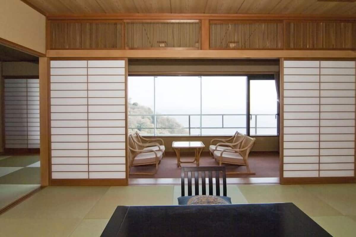 A traditional Japanese room with tatami flooring, a low table, two chairs, and a view through large windows.