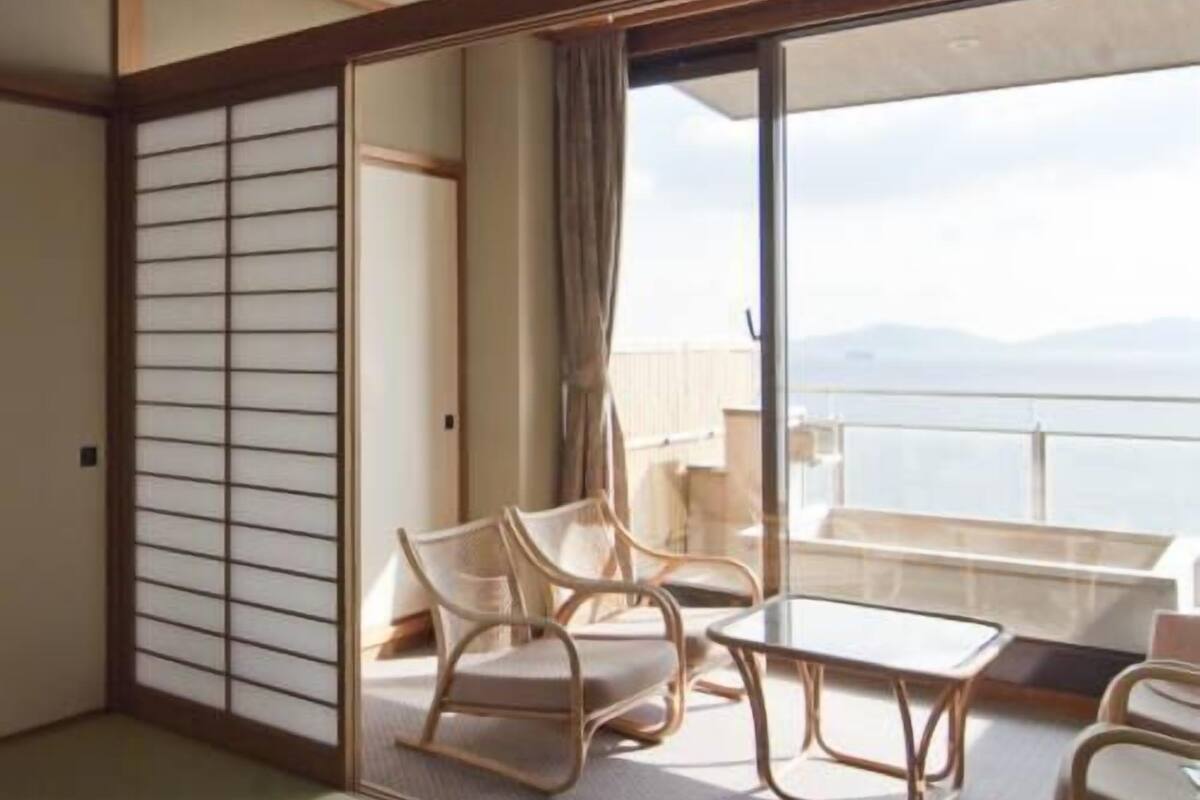 A room with tatami flooring, a sliding shoji screen, a balcony with a view, and a table with chairs.