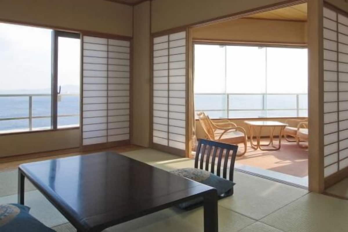 A room with tatami flooring, a sliding door leading to a balcony with a view of the sea, and a table with chairs.