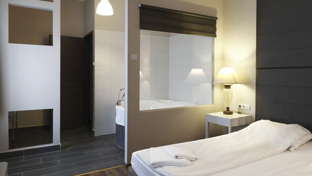 Habitación de hotel con cama, baño con lavamanos y espejo, mesita de noche con lámpara y pared de color oscuro.