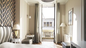Una habitación de hotel con cama, mesitas de noche, una pequeña mesa con lámpara, un banco y vistas al balcón exterior.