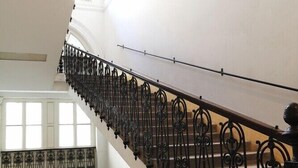 Una majestuosa escalera con barandillas negras ricamente decoradas, paredes blancas y grandes ventanales que dejan entrar la luz natural.