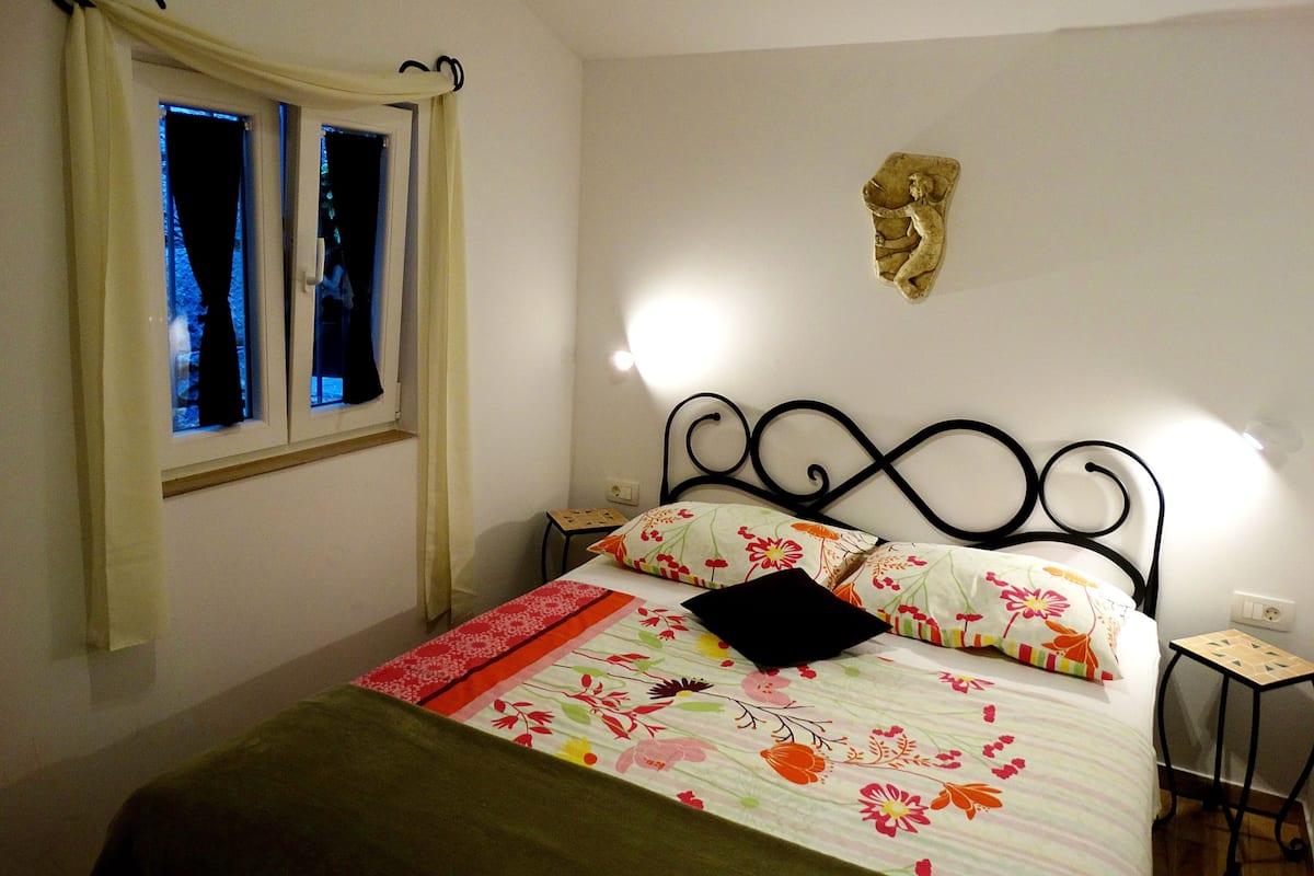 Une chambre à coucher avec un lit, une fenêtre avec des rideaux, une lampe fixée au mur et une petite table de chevet.