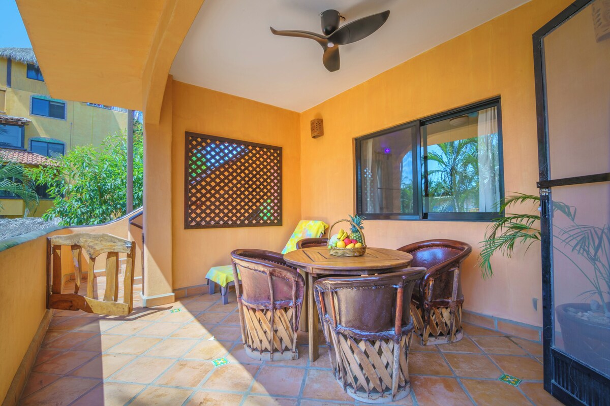 Un patio con una mesa y sillas, un ventilador de techo, una ventana con vistas a palmeras y un panel decorativo en la pared.