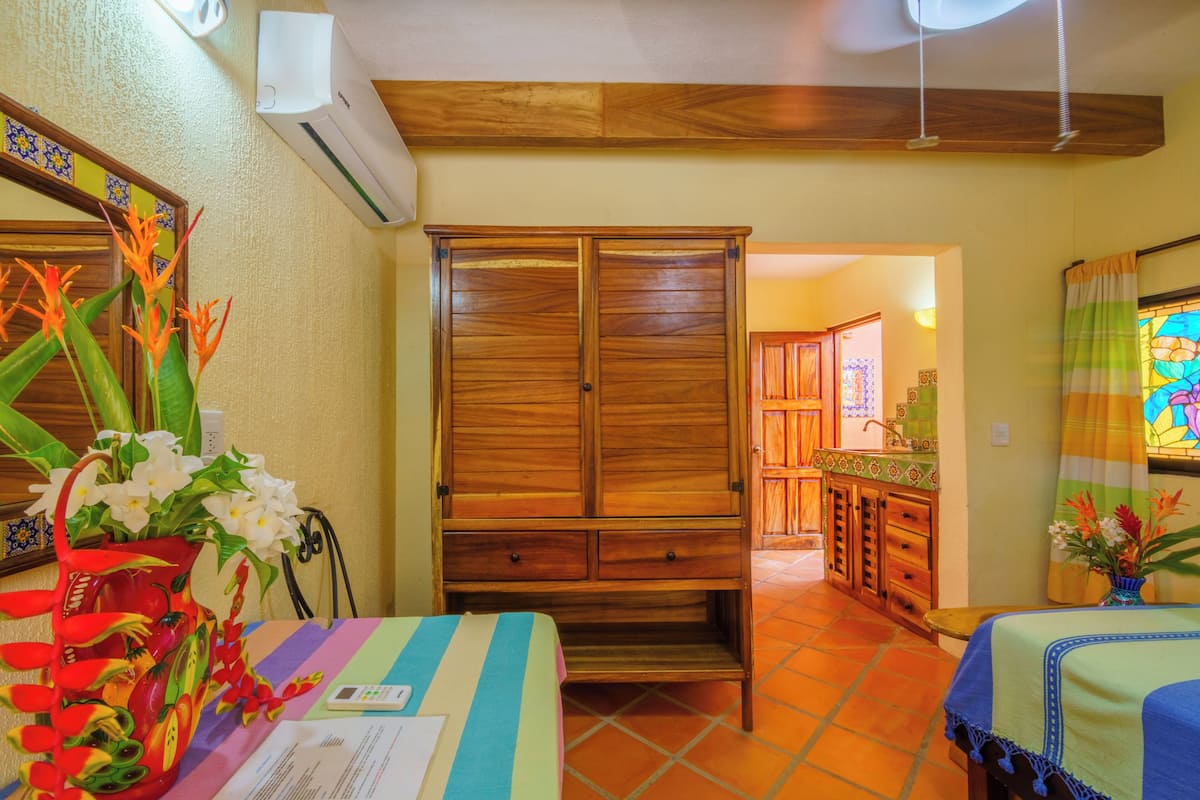 Una habitación con un armario de madera, una cama con colcha a rayas, una mesa con un jarrón de flores y una cocina visible a través de un vano abierto.