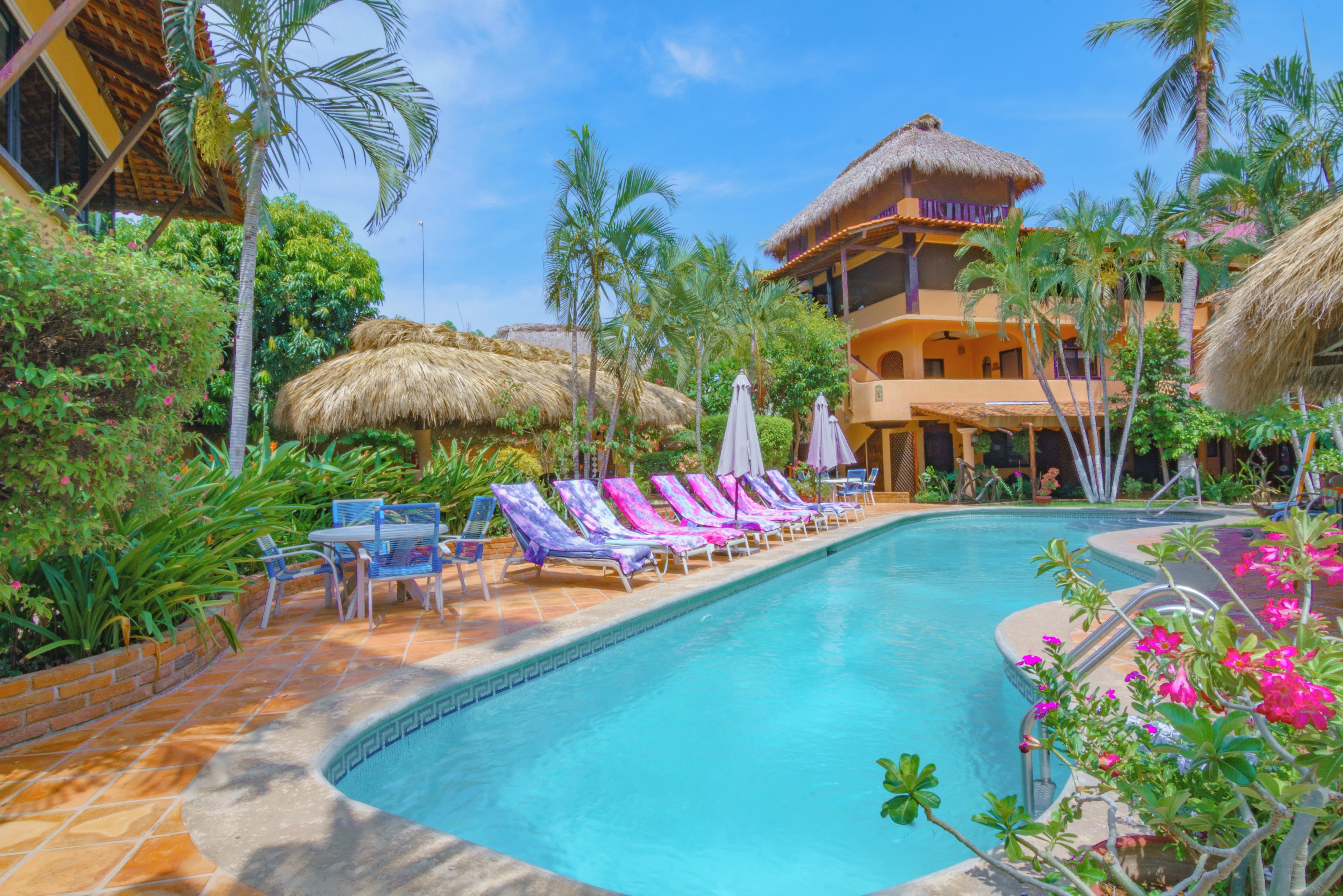 Un área de piscina con sillones de descanso y sombrillas, rodeada de vegetación tropical y un edificio con techo de paja.
