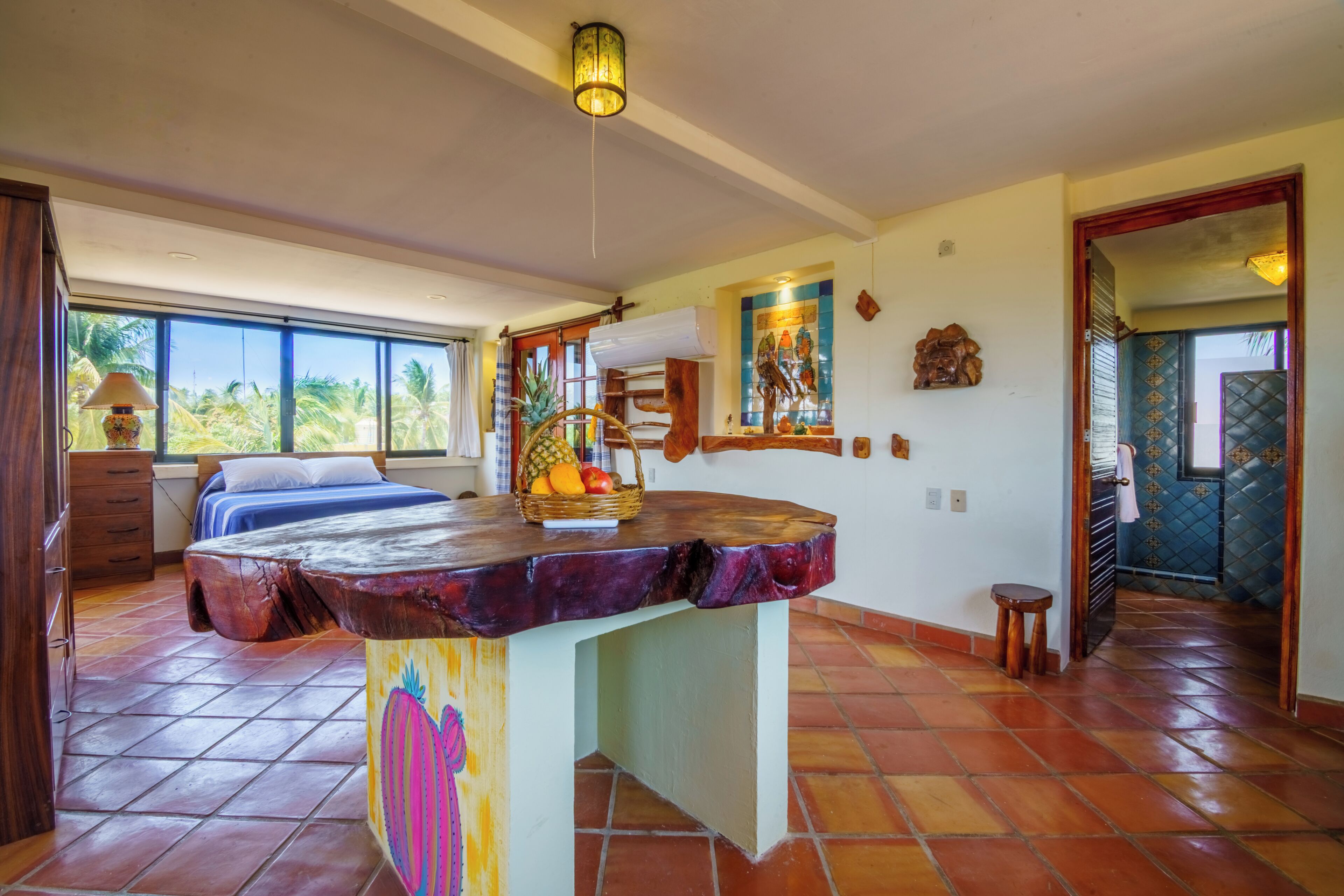 A room with a bed, a wooden table with fruit, a ceiling fan, and a view of palm trees.