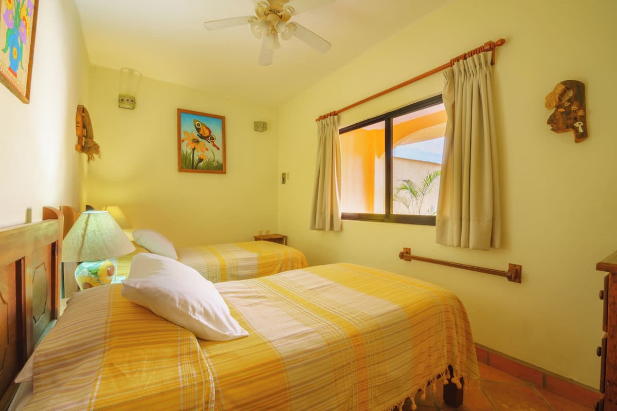 Una habitación con dos camas, ventilador de techo, ventana con cortinas, un cuadro y cabecera de madera.