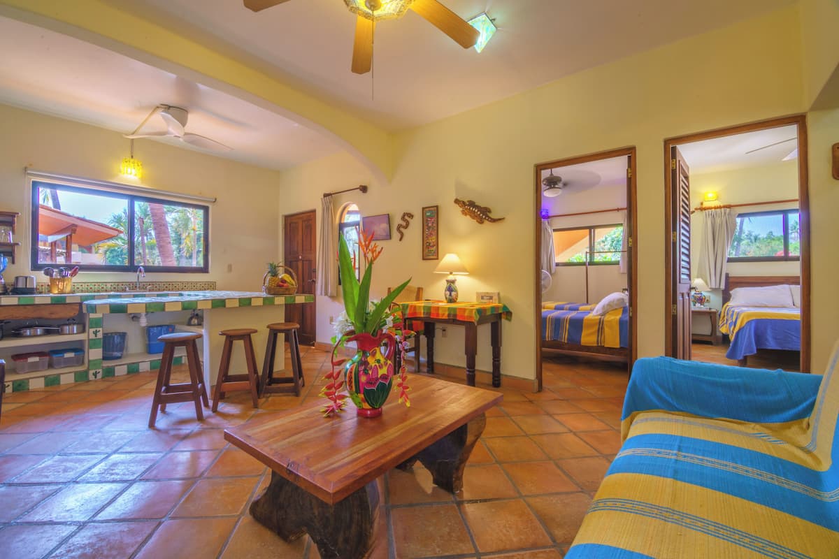 Habitación de hotel con mesa de comedor de madera, un sofá, una zona de cocina con barra y vista a palmeras.