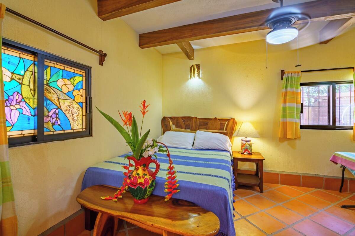 Un dormitorio con cama, mesita de noche, mesa, jarra y ventana con cortinas.