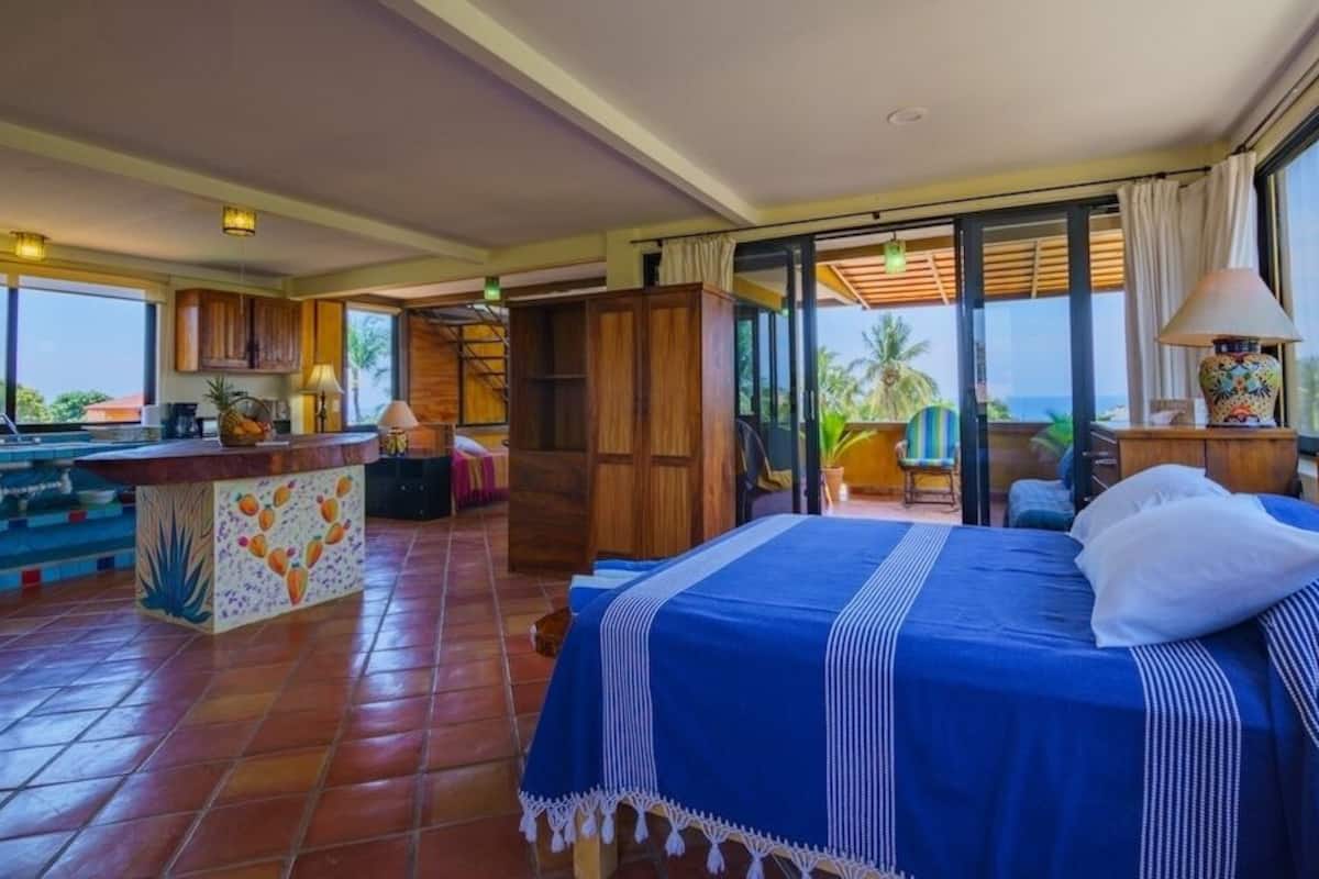 Un dormitorio con una cama grande, vista al océano, una kitchenette y un balcón con una silla.