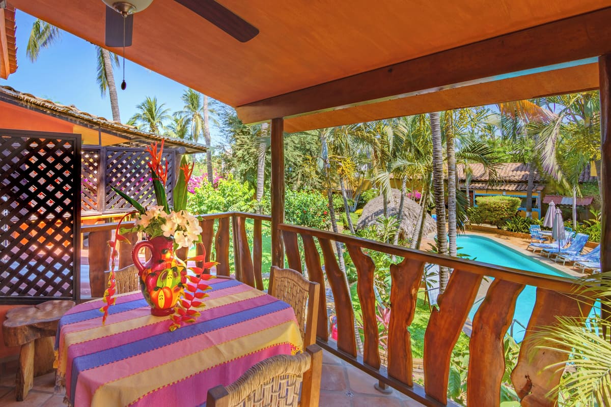 Un espacio al aire libre techado con piscina, sillas de descanso, una mesa con mantel colorido y un jarrón decorativo.