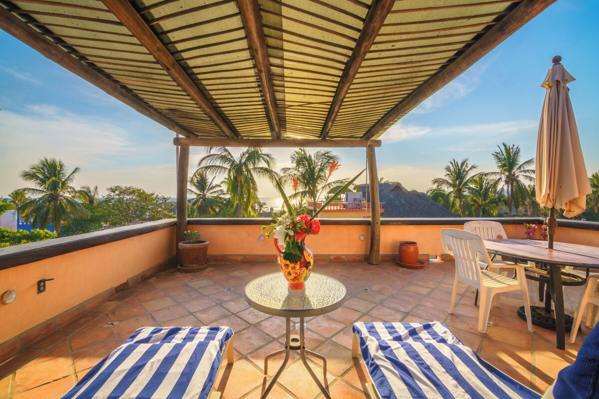 Un espacio al aire libre cubierto con una mesa, sillas, un jarrón decorativo y vistas a palmeras y montañas.