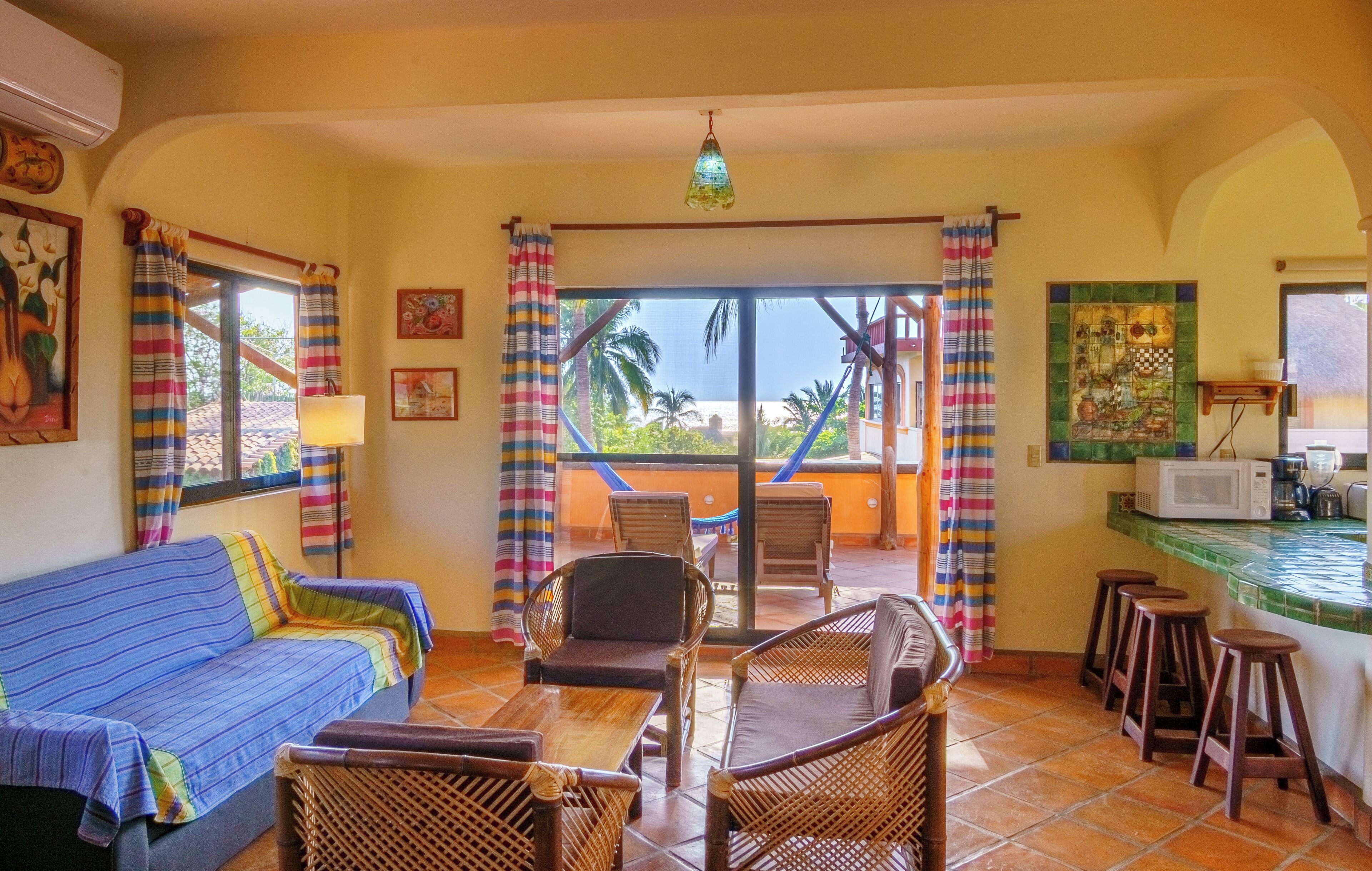 A room with a sofa, wicker chairs, a dining table, a kitchen area with a microwave, and a view of palm trees through the balcony doors.