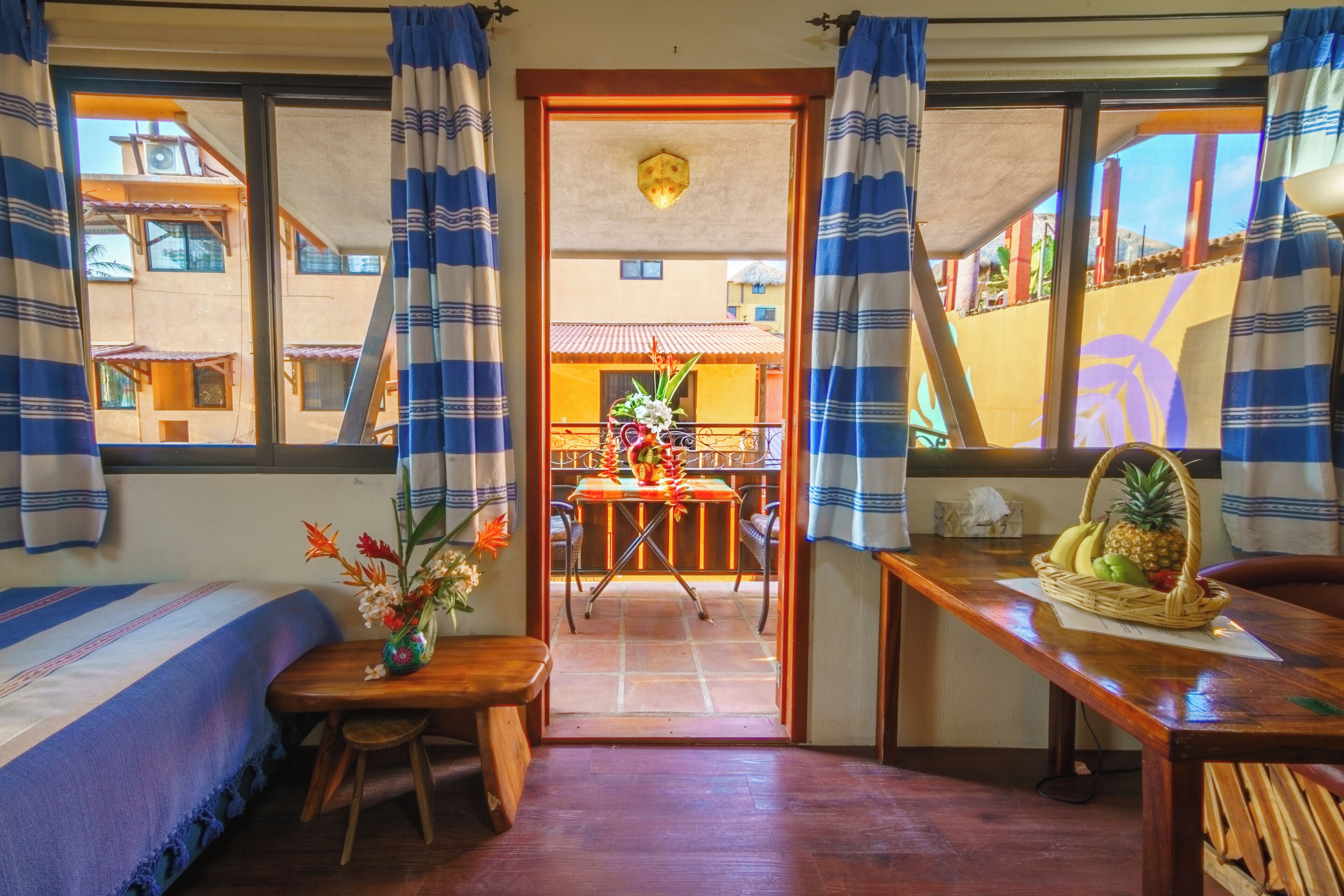 A room with a bed, a table with a fruit basket, a vase with flowers, and a view of the outside balcony.