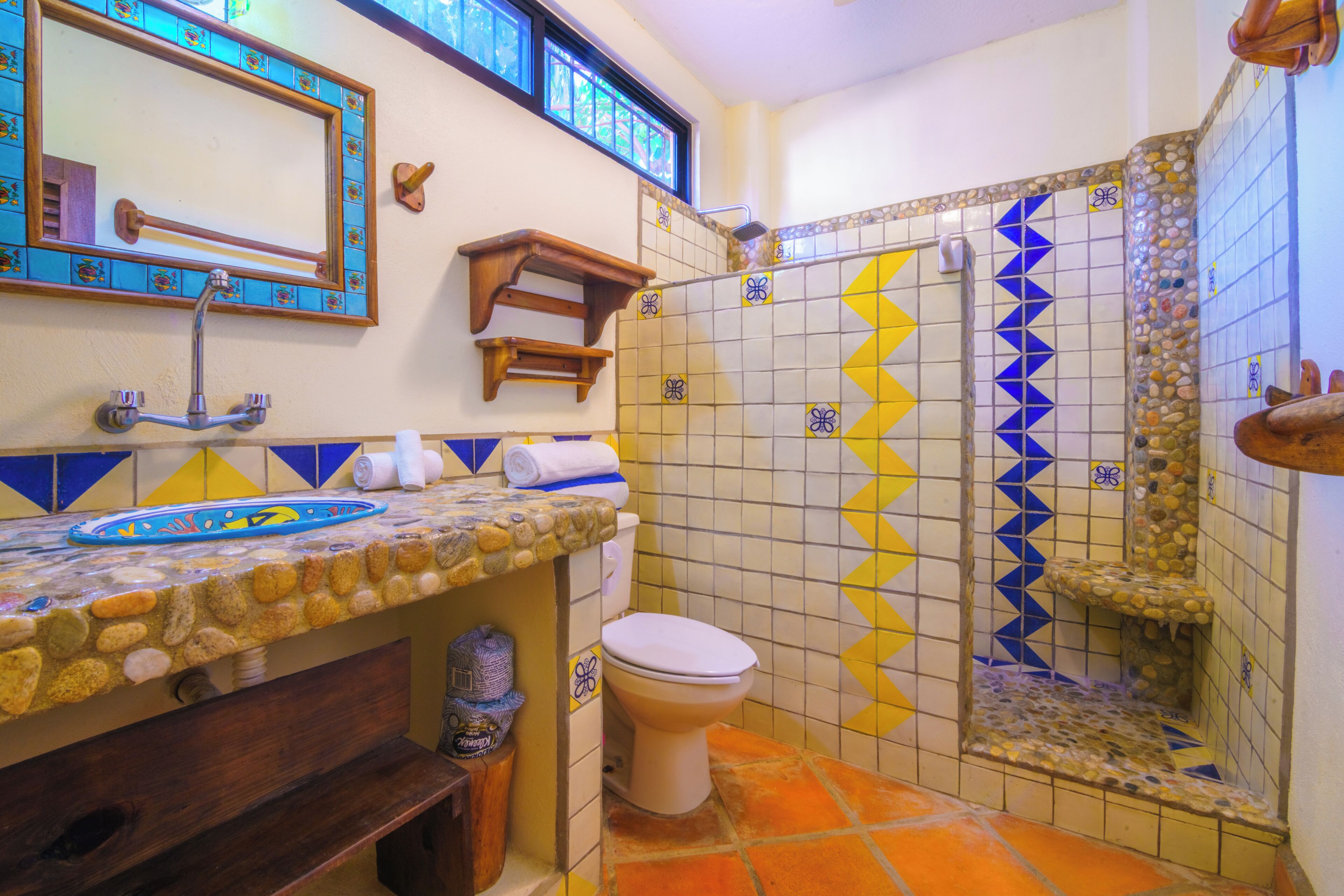 A bathroom with a stone countertop, a toilet, a shower with mosaic tiles, and a wooden shelf.