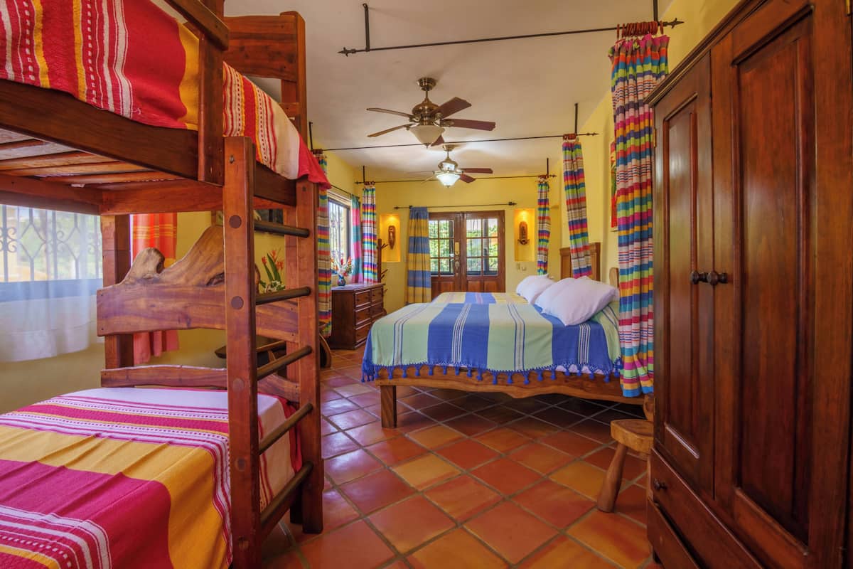 Una habitación con una cama individual, una litera, un armario de madera, un ventilador de techo y una ventana con cortinas.