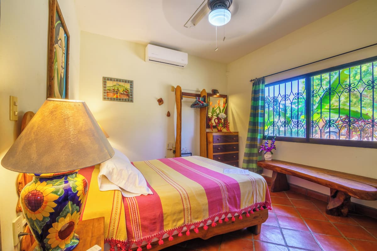 Una habitación con una cama, una lámpara, un banco, una ventana con vistas y un equipo de aire acondicionado.