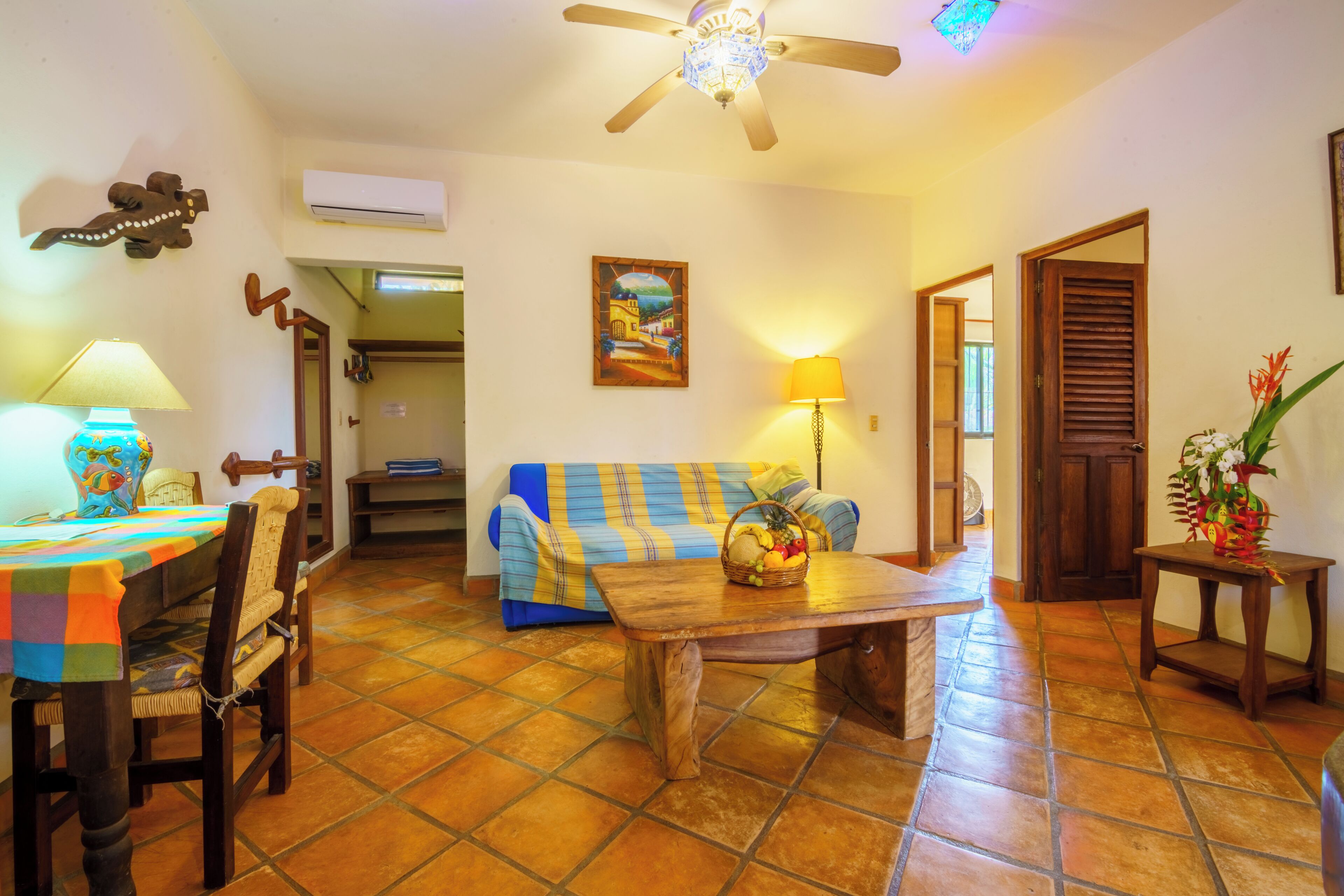 A living room with a wooden coffee table, a blue and yellow sofa, a dining table with chairs, a ceiling fan, and a wall-mounted air conditioner.