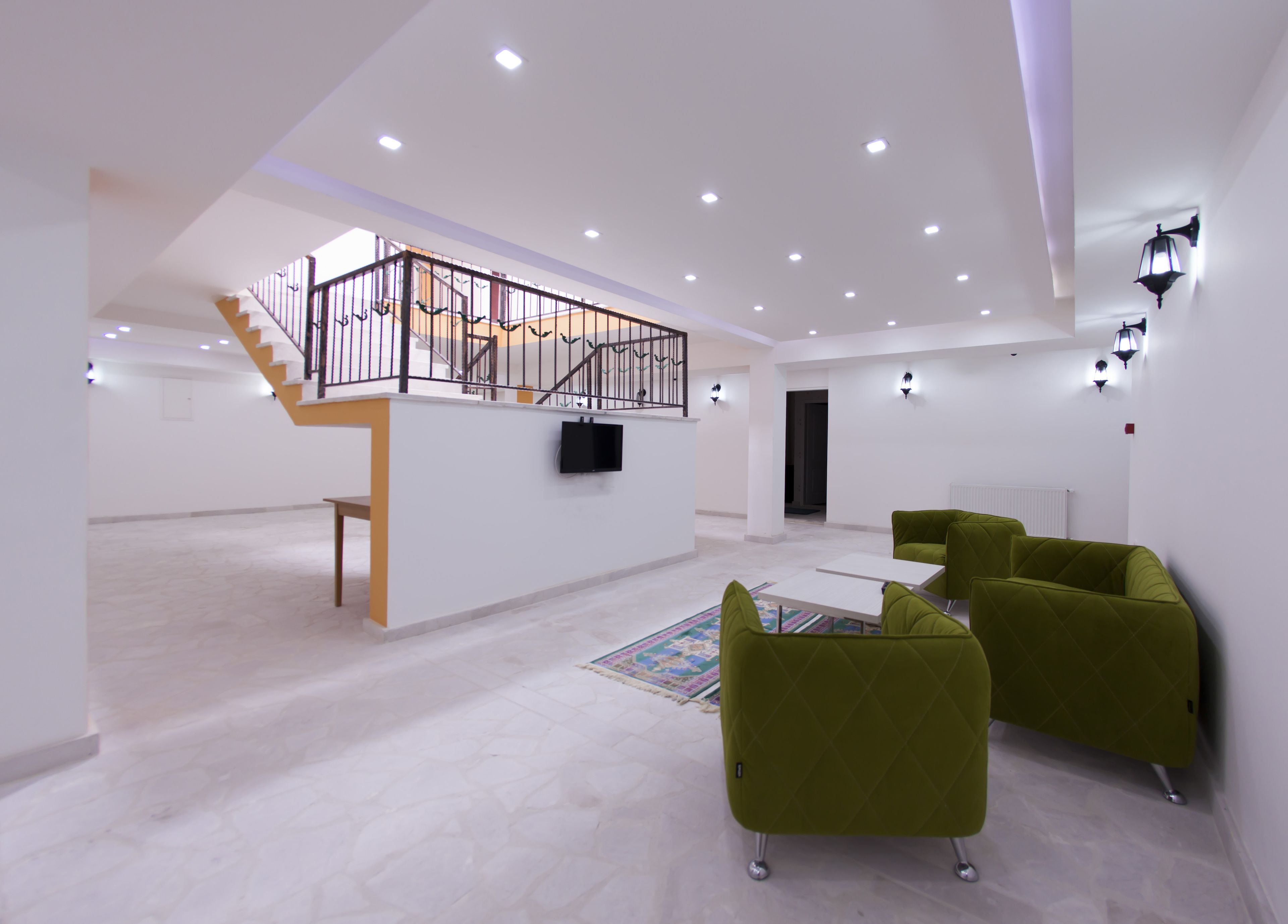 A spacious living area with a staircase leading to an upper level, a flat-screen TV mounted on a white wall, and green upholstered chairs.