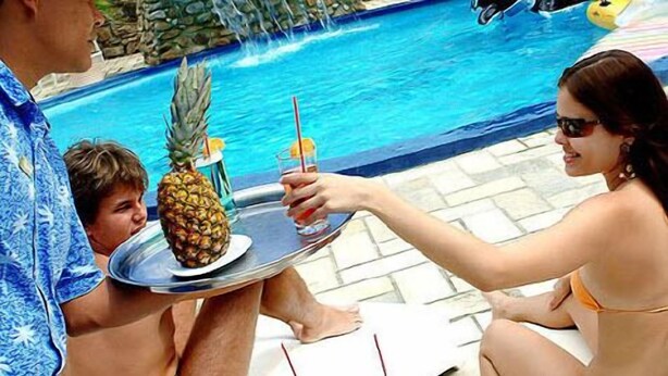En miljö vid poolkanten med en person som håller i ett bricka med en ananas och en drink.