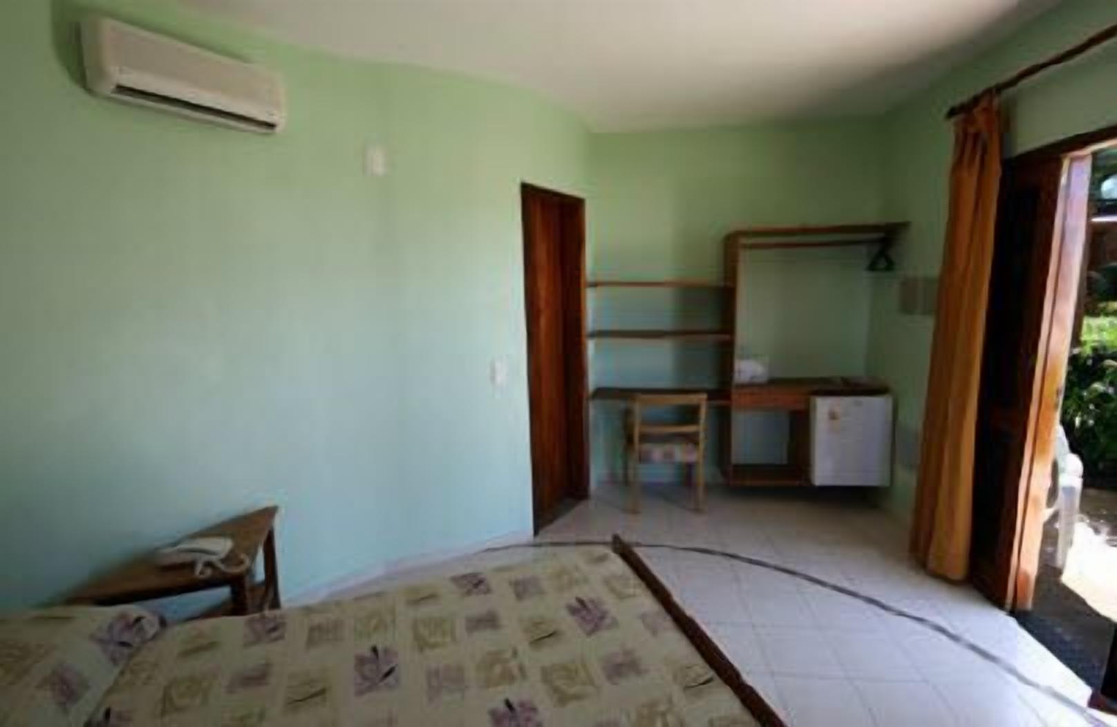 Quarto com cama, escrivaninha e cadeira. Tem uma janela com cortinas, um radiador e uma porta.