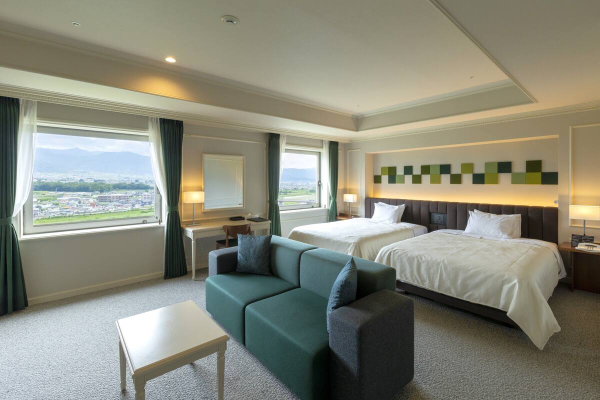 A hotel room with two beds, a sofa, a desk, and a view of a cityscape.