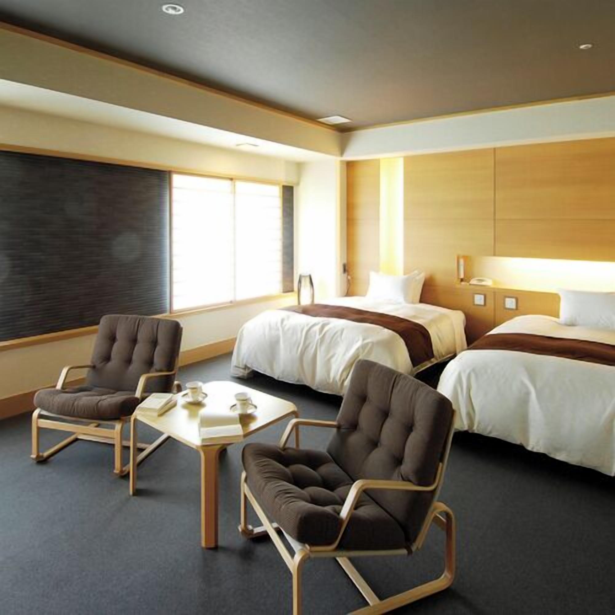 A hotel room with two beds, a desk, two armchairs, and a large window.