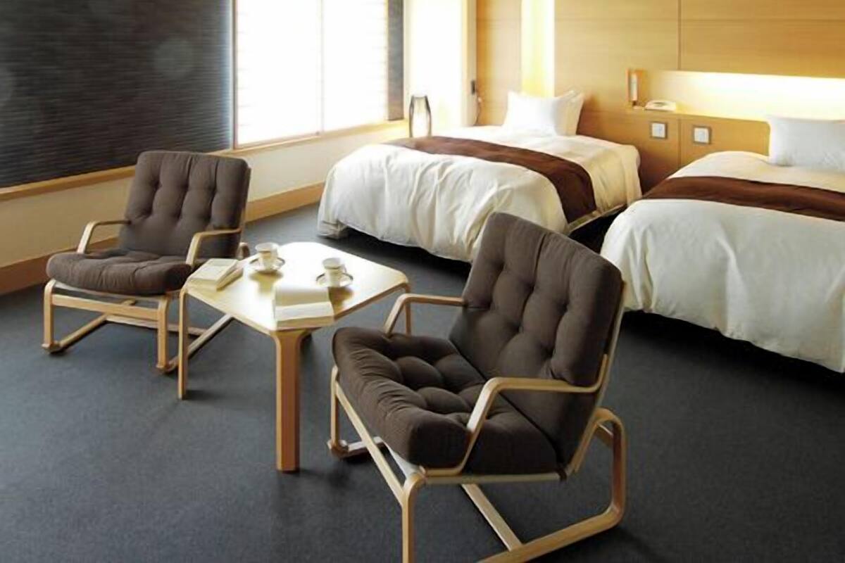 A hotel room with two beds, a desk, two armchairs, and a large window.