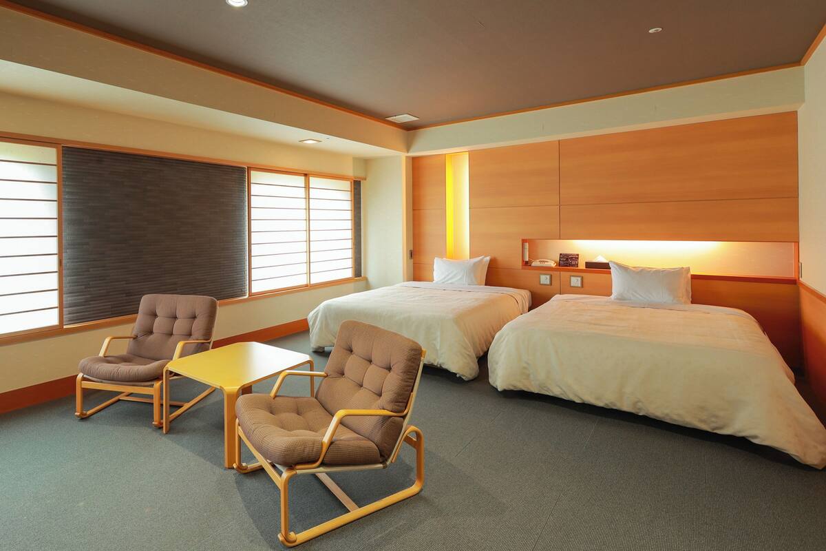 A hotel room with two beds, a small table, two armchairs, a window with blinds, and a wooden headboard.