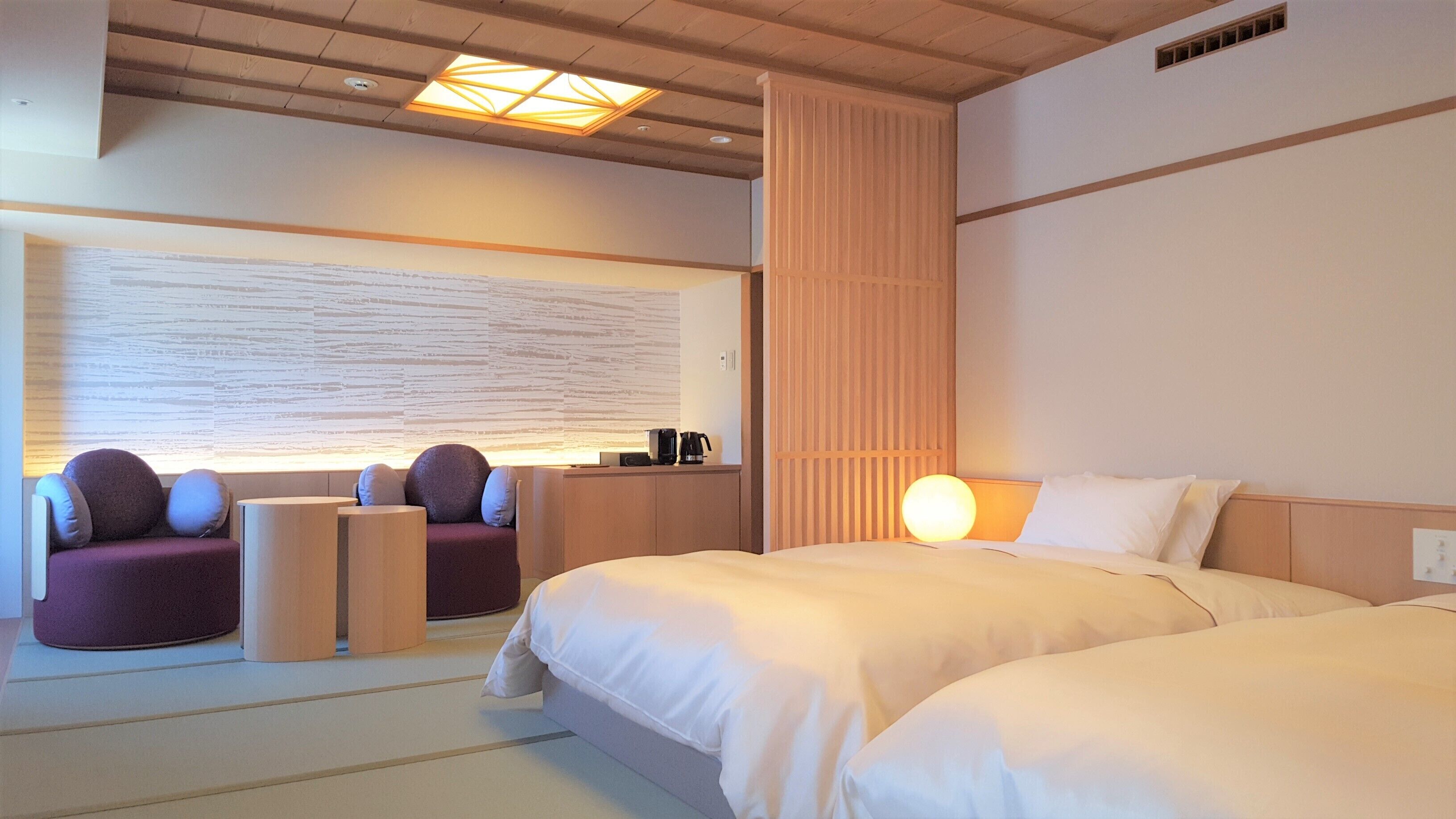 A hotel room with two beds, a seating area with a sofa and chairs, a wall-mounted desk with a coffee maker, and a large abstract wall art piece.
