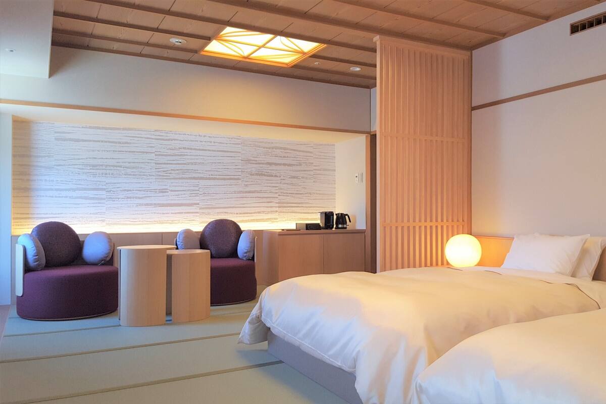 A hotel room with two beds, a seating area with a sofa and chairs, a wall-mounted desk with a coffee maker, and a large abstract wall art piece.