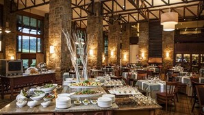 A spacious dining area with high ceilings and exposed beams, featuring a buffet setup with various dishes and a central decorative tree. - Drakensberg Sun (Cathkin Park)