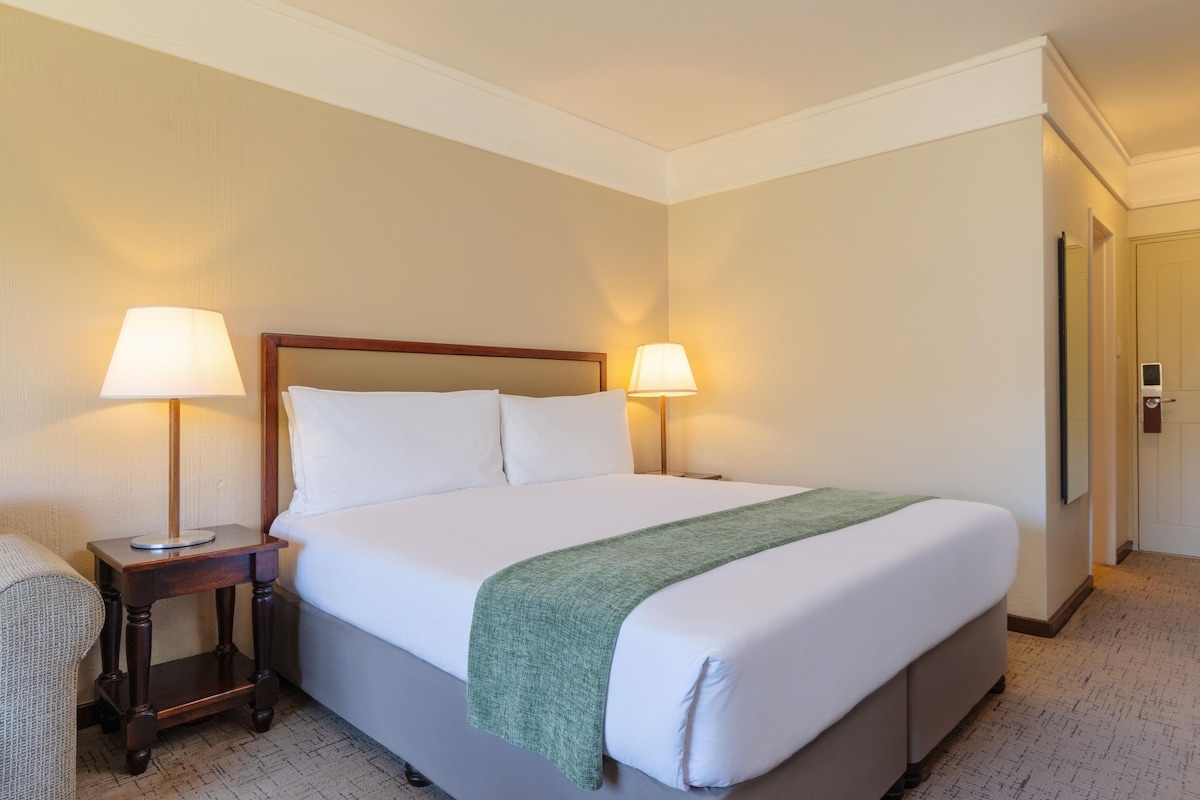 A hotel room with a bed, two bedside lamps, a green throw blanket, and a door.