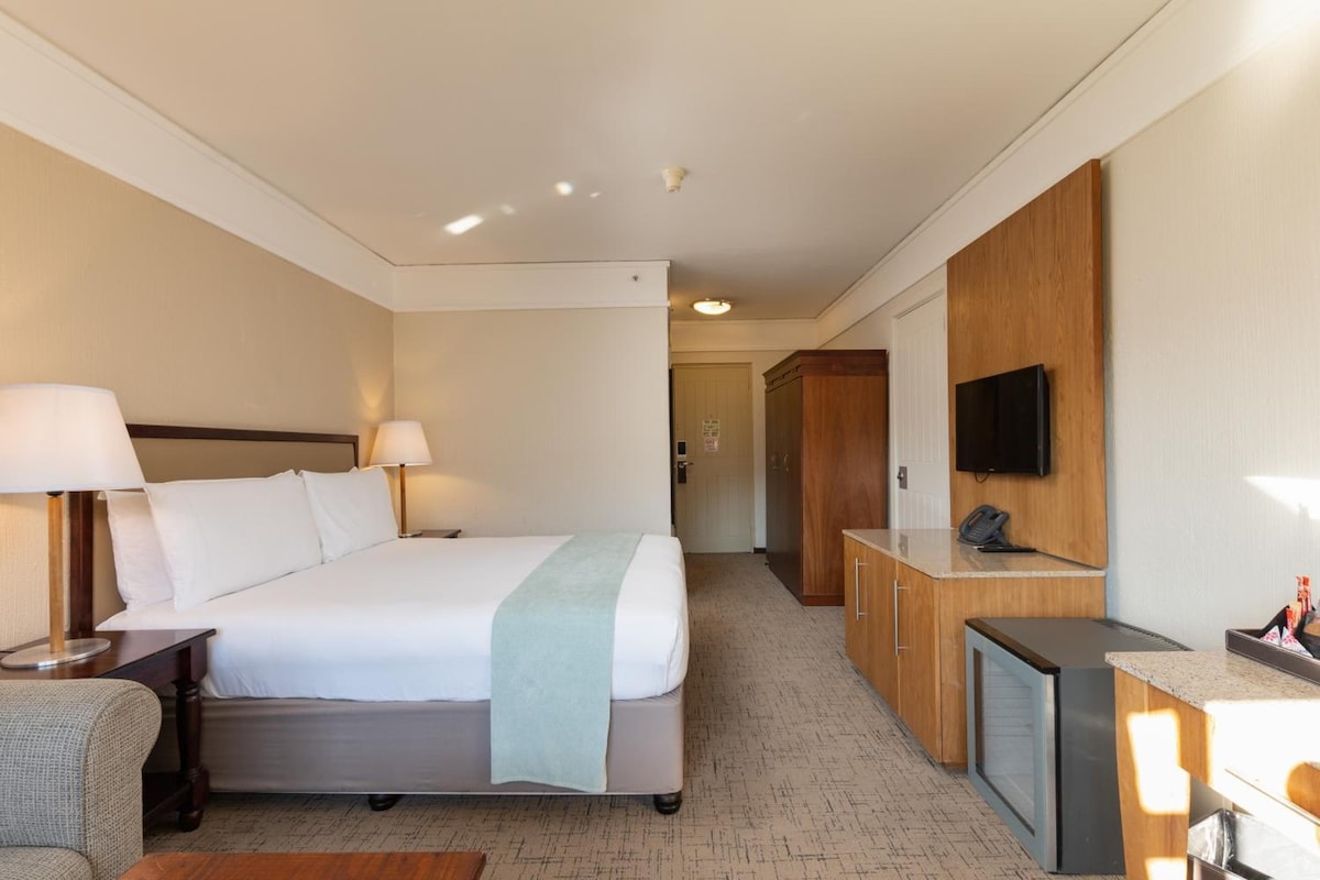 A hotel room with a large bed, bedside lamps, a television, a desk with a telephone, and a wardrobe.