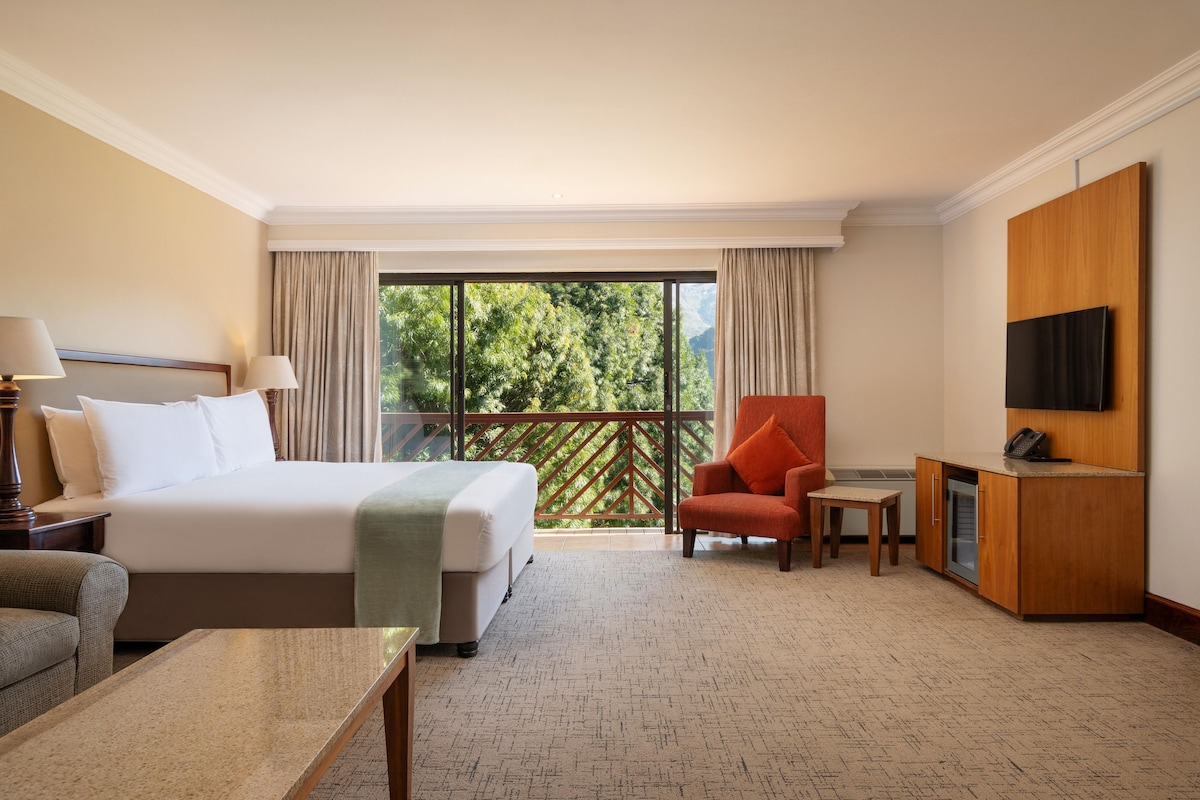 A hotel room with a bed, a desk, a chair, a television, and a balcony with a view of trees and mountains.