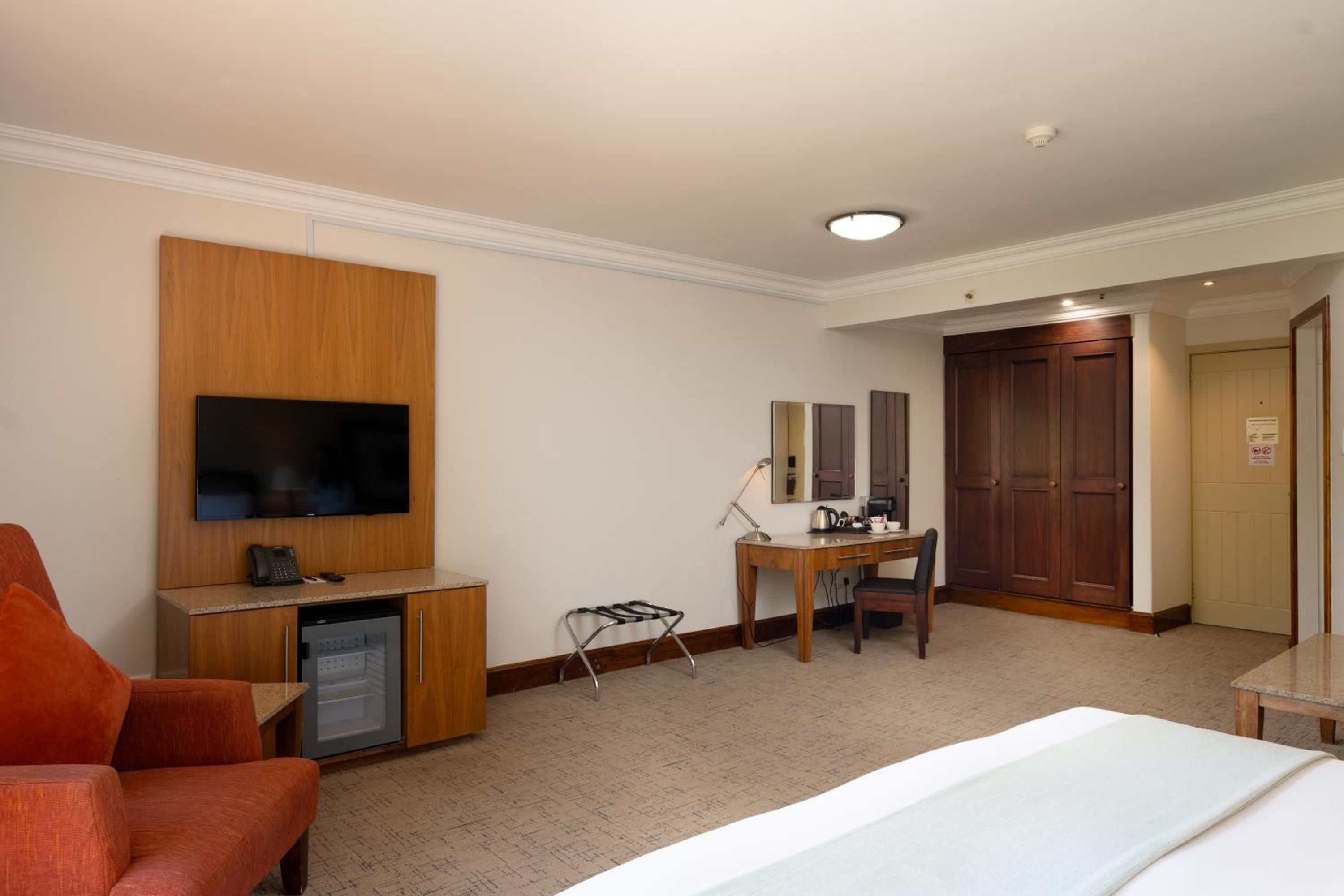 A hotel room with a bed, a desk with a chair, a television, a small refrigerator, and a wardrobe.