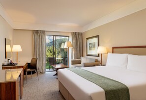 Quarto de hotel com uma cama, duas cadeiras, uma escrivaninha e vista para o exterior.