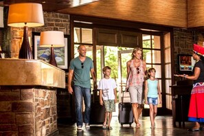 A family walking through a lobby with a stone wall, a wooden ceiling, and a large window. - Drakensberg Sun (Cathkin Park)