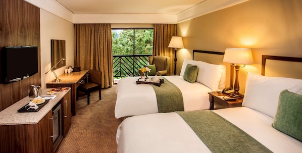 A hotel room with two beds, a desk, a TV, and a balcony with a view of greenery. - Drakensberg Sun (Cathkin Park)