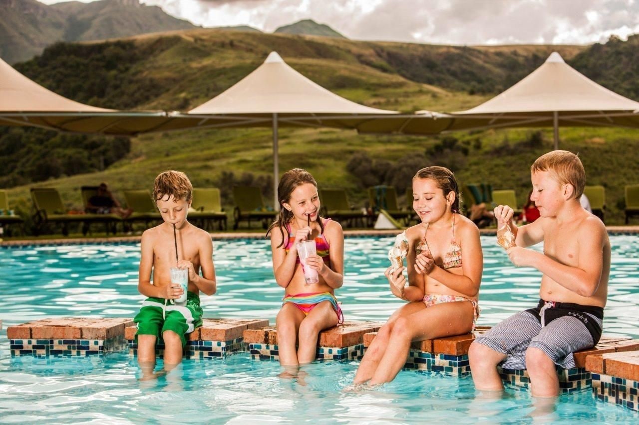 A poolside setting with people sitting on the edge, a tiled pool, and a mountainous backdrop.