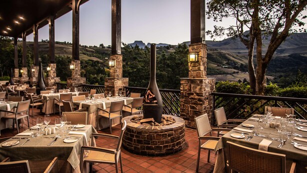An outdoor dining area with tables set for a meal, stone pillars, and a fire feature. - Drakensberg Sun (Cathkin Park)