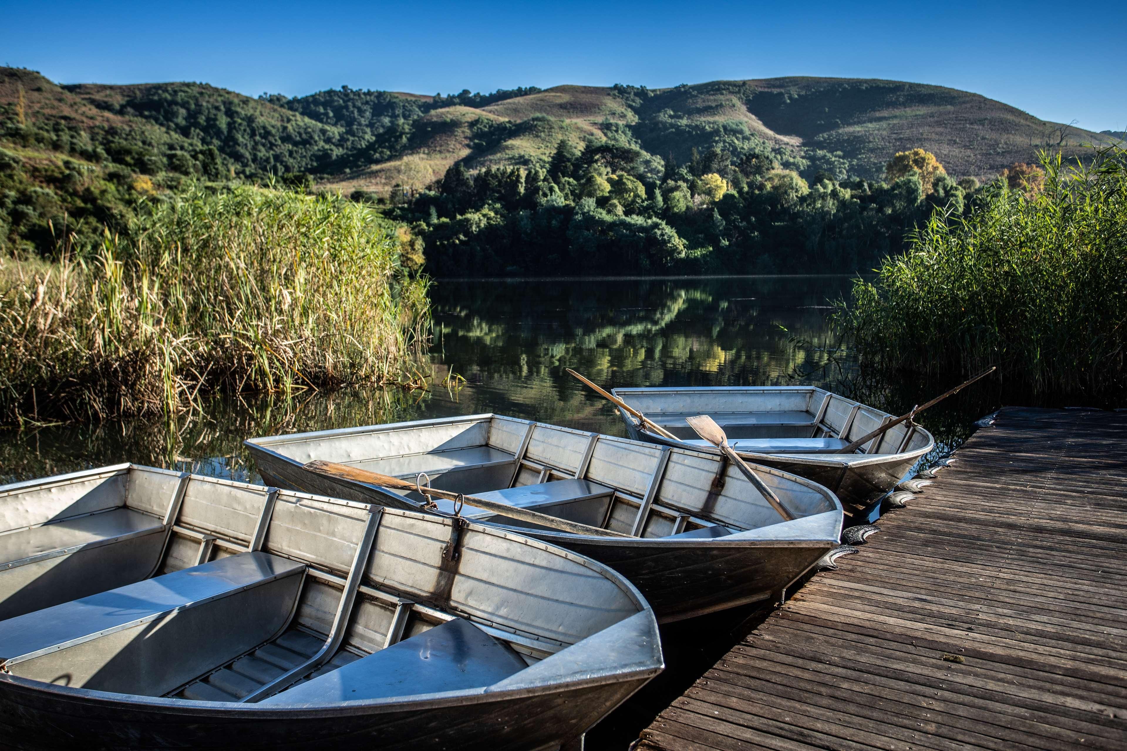 Een houten steiger met verschillende kleine boten afgemeerd aan een rustig meer, omringd door weelderige begroeiing en heuvels op de achtergrond.