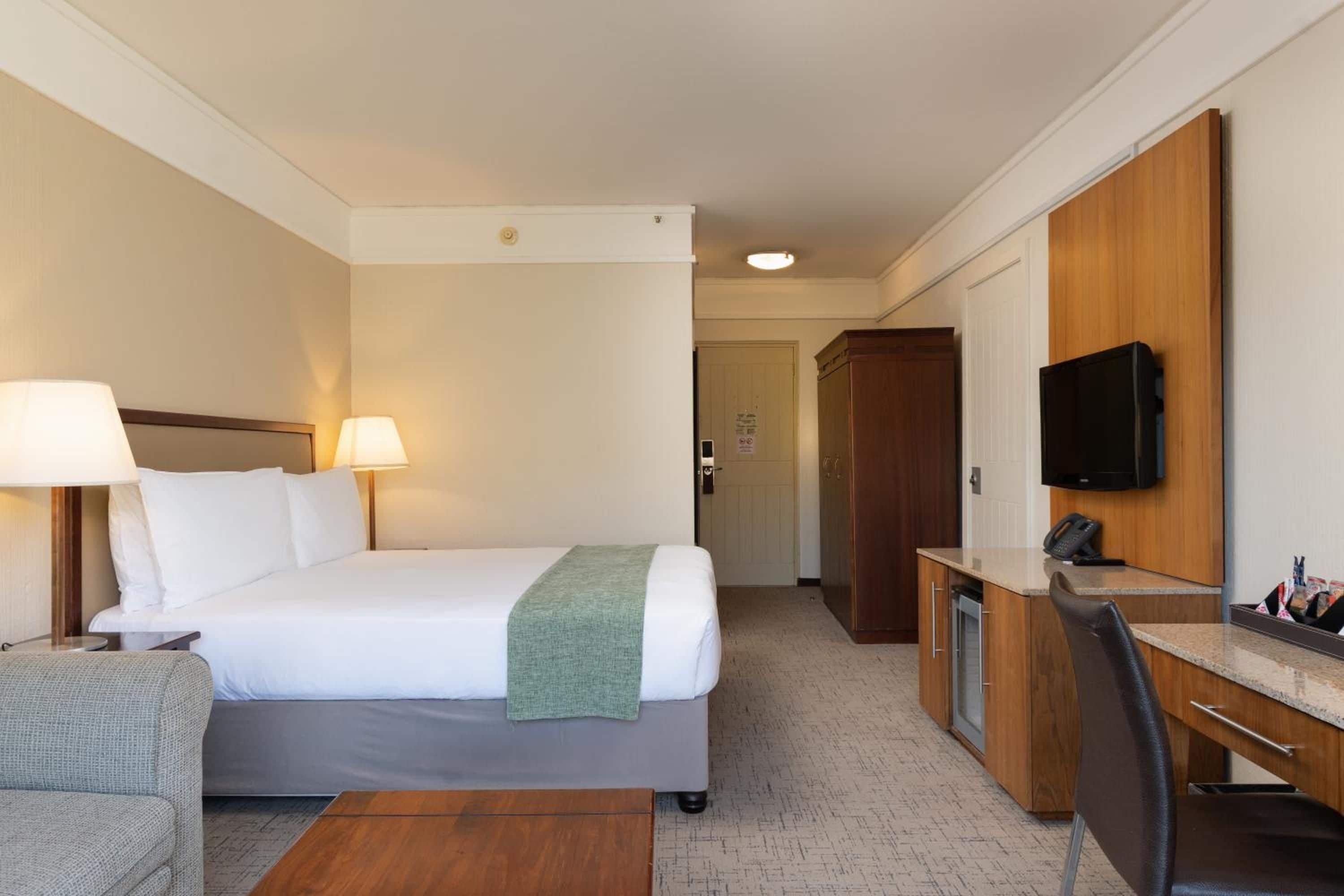 A hotel room with a bed, bedside lamps, a desk with a chair, a television, and a wardrobe.