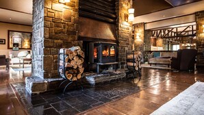 A stone fireplace with a log fire, surrounded by wooden furniture and a stone wall. - Drakensberg Sun (Cathkin Park)