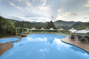 Piscina com borda curva, rodeada por espreguiçadeiras e guarda-sóis, com colinas verdes e céu nublado ao fundo.